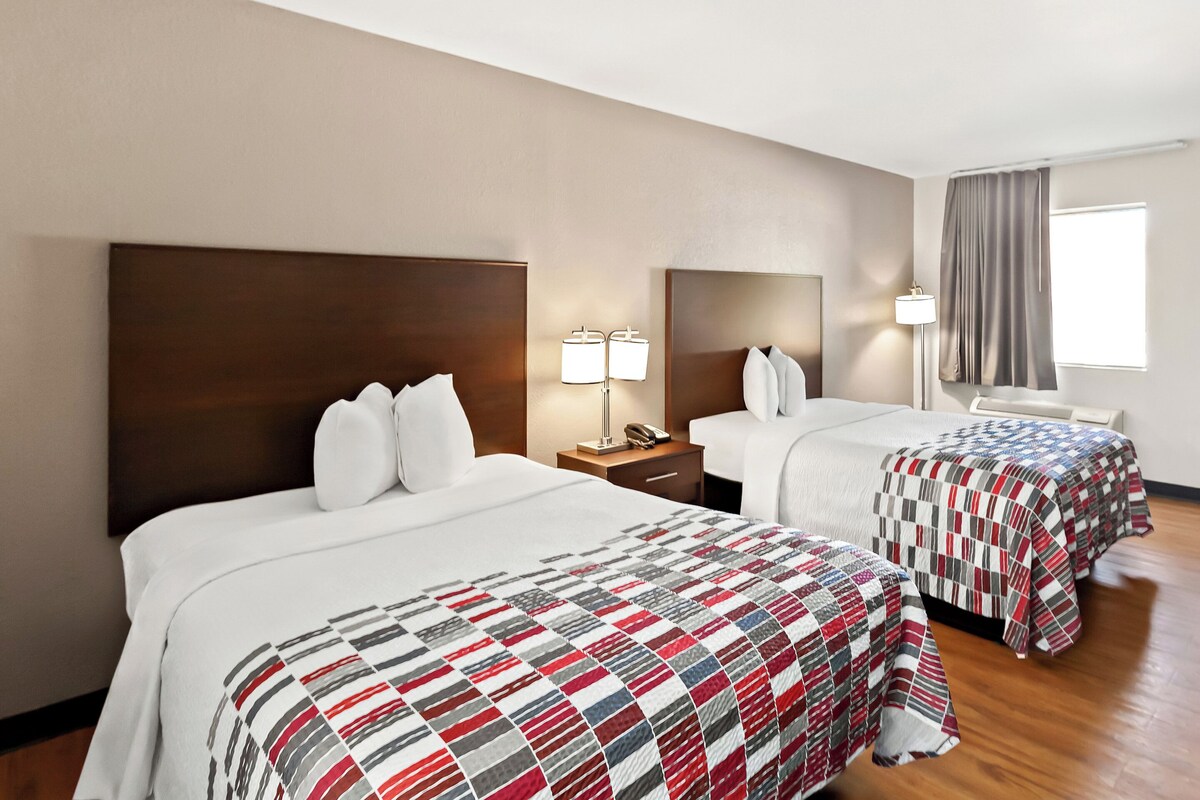 A hotel room with two beds, a nightstand, a lamp, and a window with curtains.