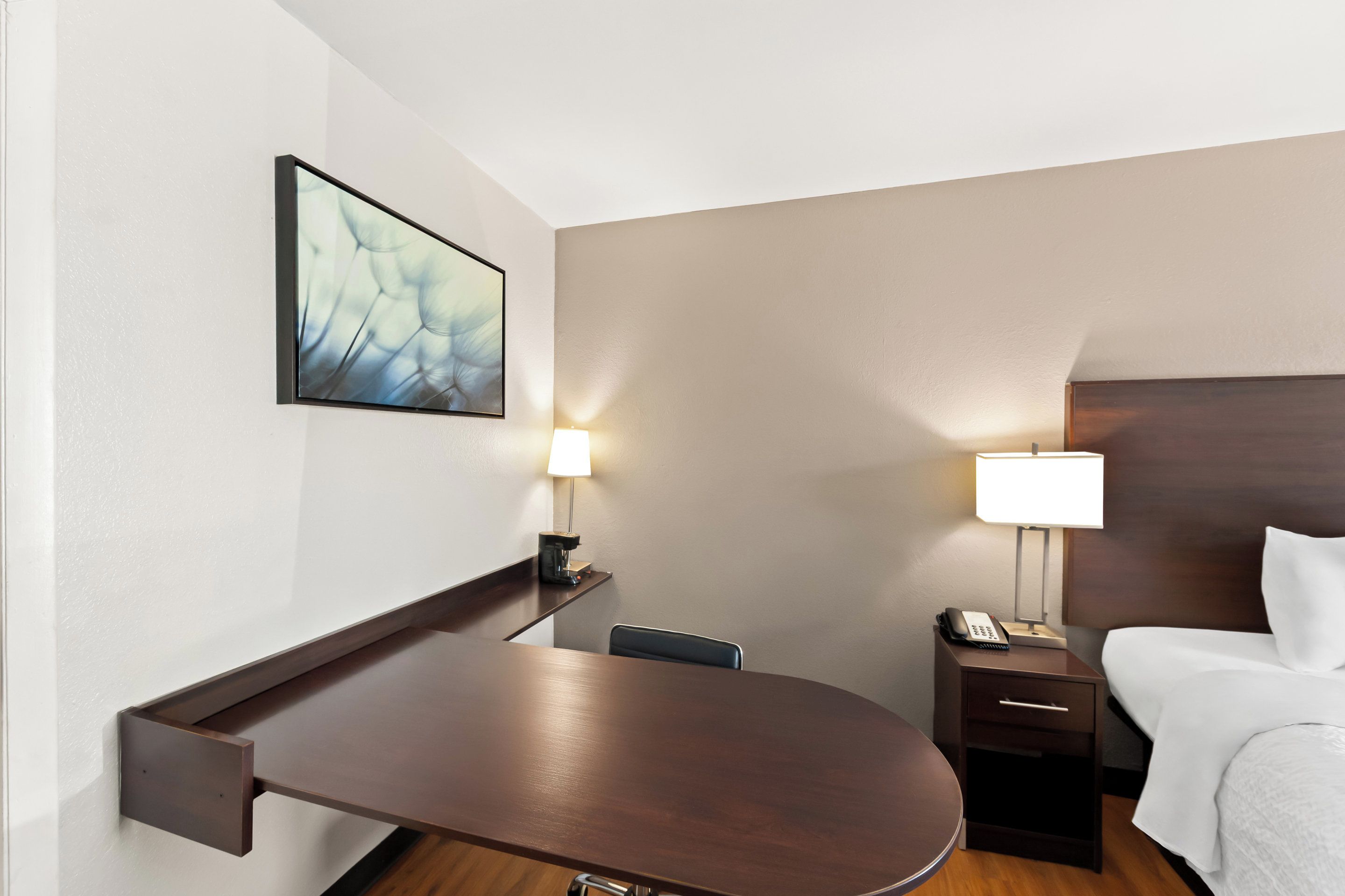 A hotel room with a wooden desk, a bed, a nightstand with a lamp, and a wall-mounted picture.