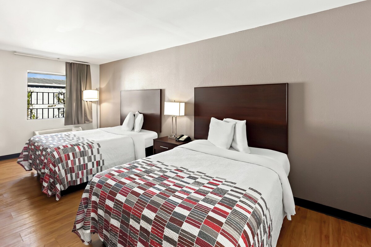 A hotel room with two beds, a window with curtains, and a nightstand with a lamp.