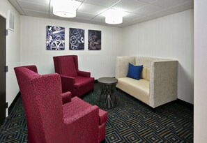 Habitación de hotel con un sofá rojo estampado, un sofá beige con una almohada azul y un sillón rojo estampado. Hay tres obras de arte colgadas en la pared que representan engranajes mecánicos.