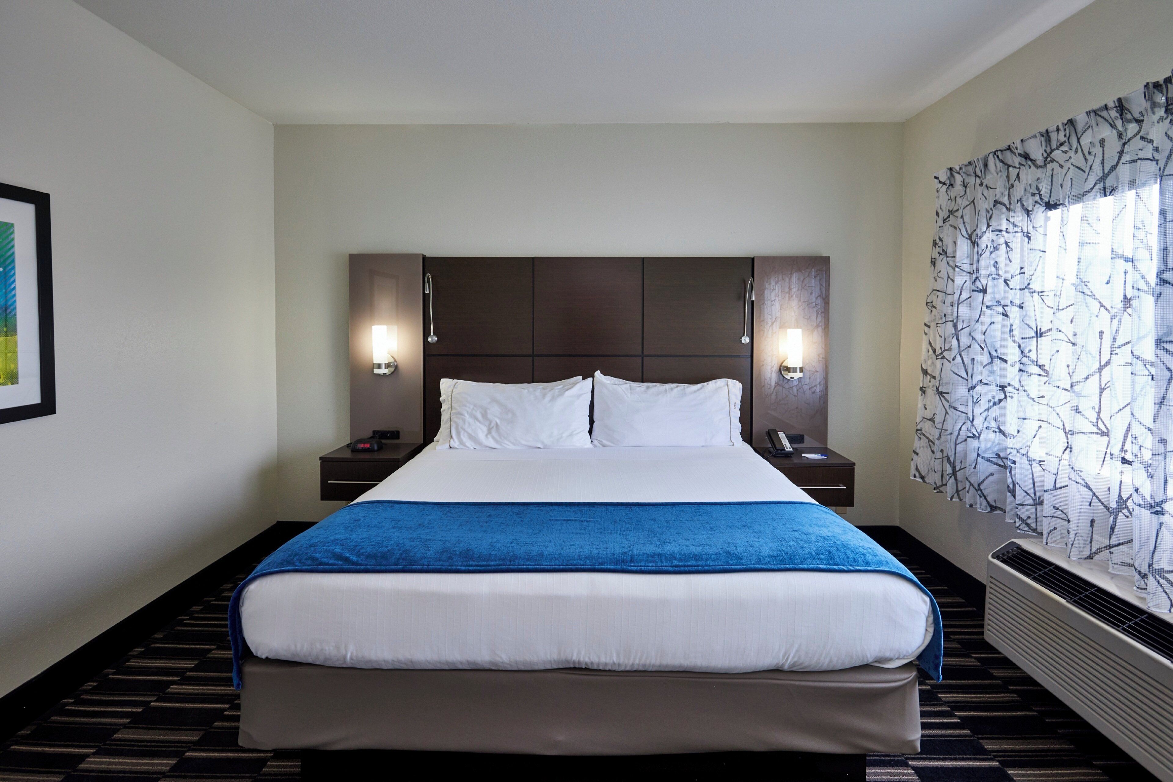 A hotel room with a large bed, two bedside tables, wall-mounted lamps, a window with patterned curtains, and a radiator.