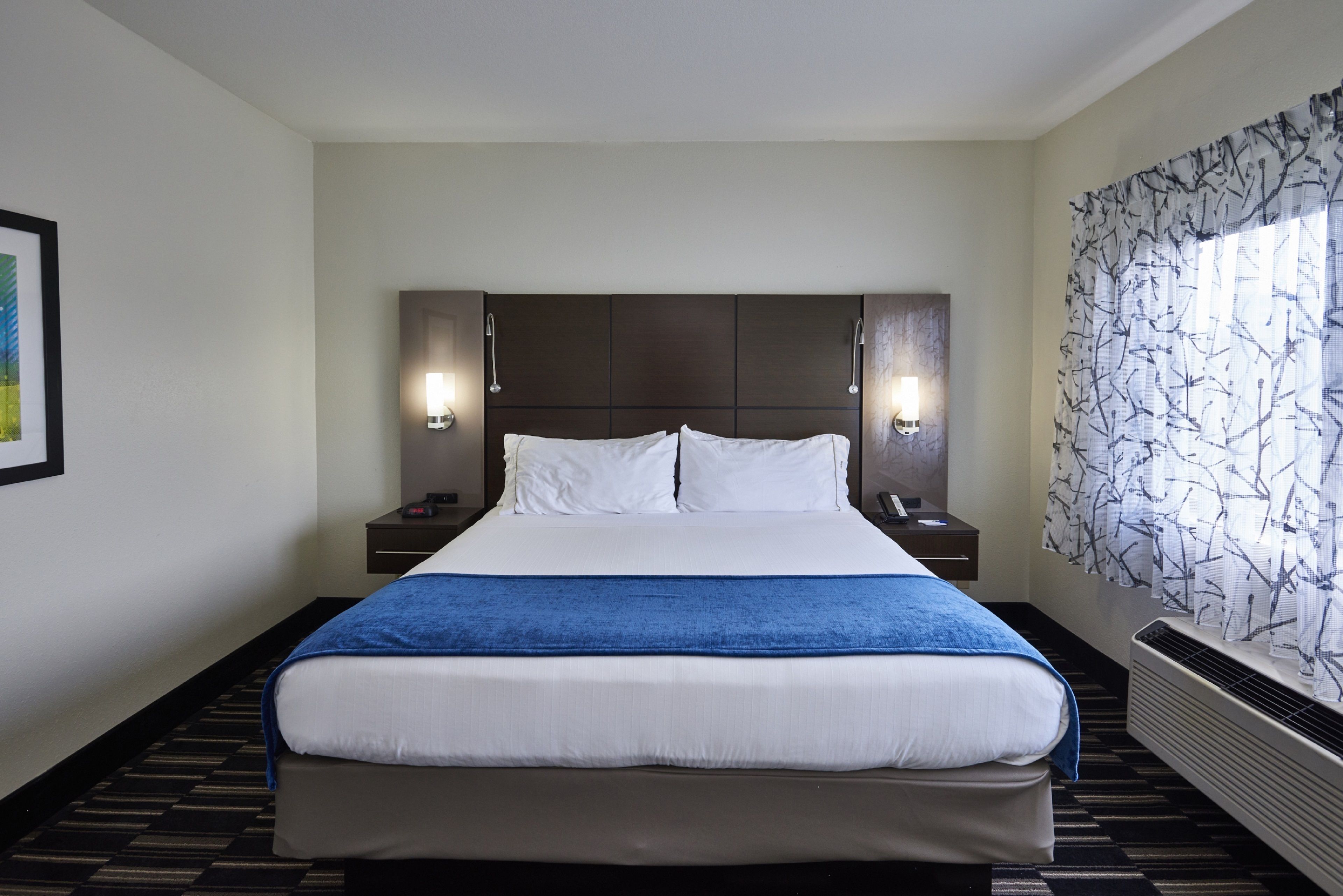 Una camera d'albergo con un letto grande, due comodini, lampade a parete e una finestra con tende a motivi, oltre a un quadro appeso al muro.