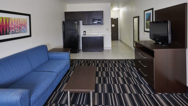 Una sala de estar moderna con un sofá azul, una mesa de centro, un televisor de pantalla plana y una zona de cocina con armarios oscuros y un microondas.