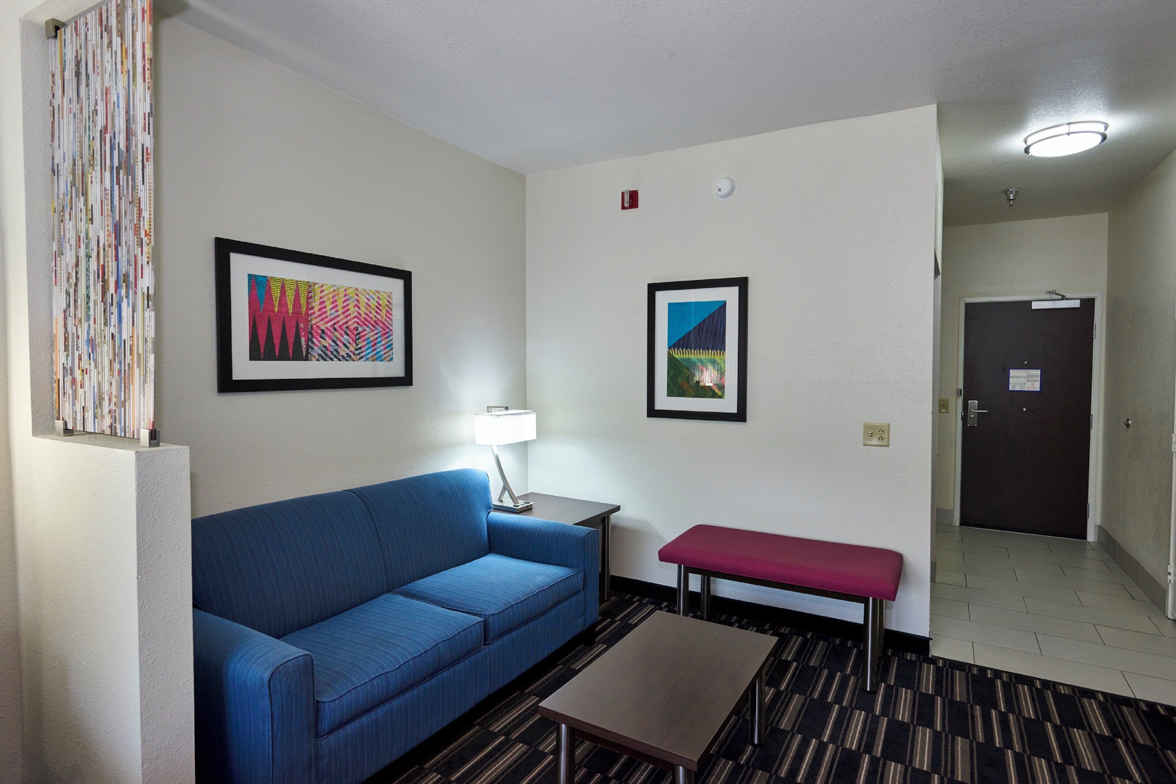 A hotel room with a blue sofa, a small table, a bench, and two framed artworks on the wall.