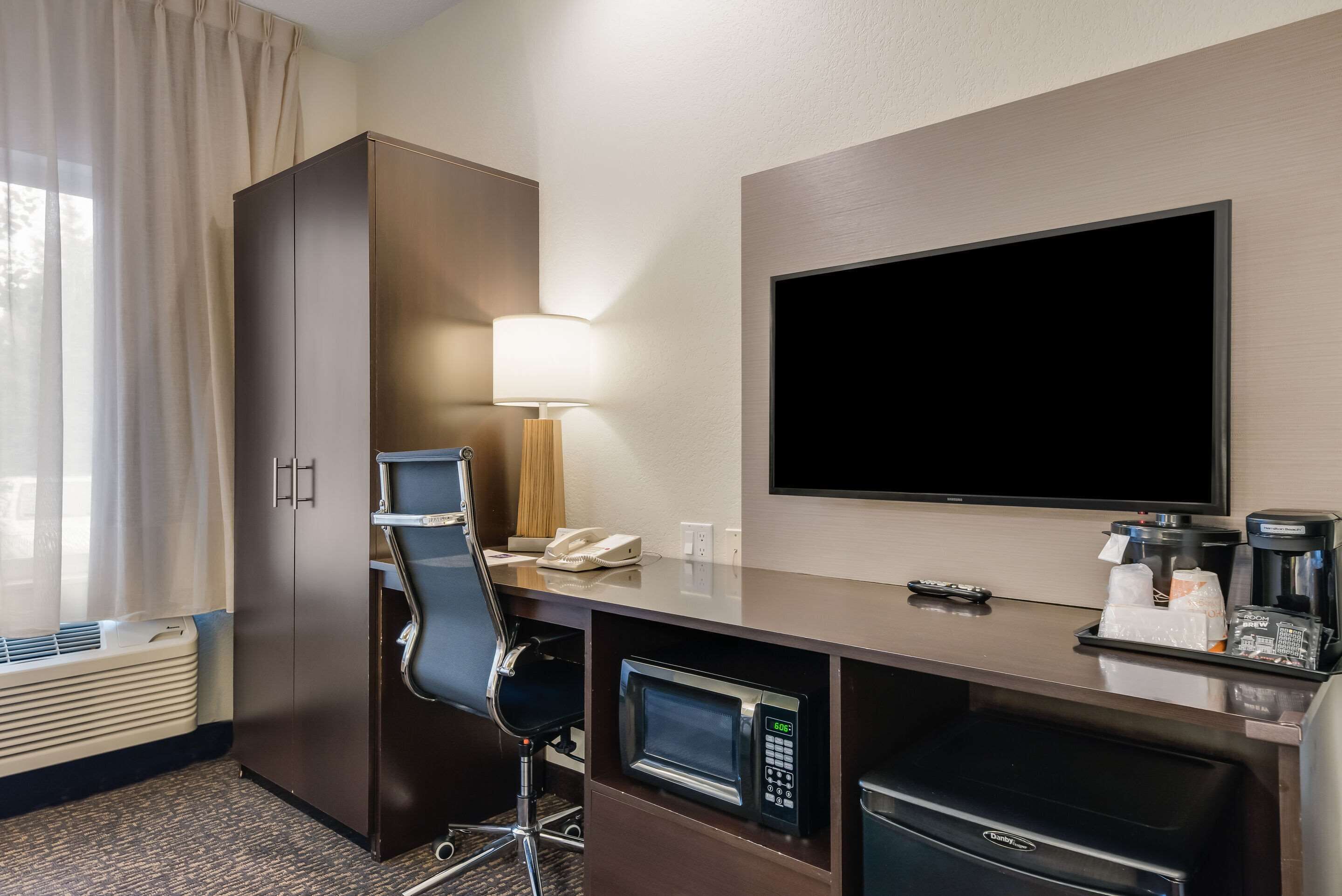 A hotel room with a desk, a microwave, a coffee maker, a TV, a chair, and a wardrobe.
