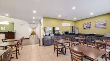 Uma sala de jantar limpa e bem iluminada, com várias mesas e cadeiras, uma área de cozinha com eletrodomésticos e uma saída visível.