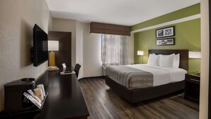Una habitació d'hotel amb un llit, una taula de treball, una cadira, un televisor, una finestra amb cortines i una paret amb quadres.
