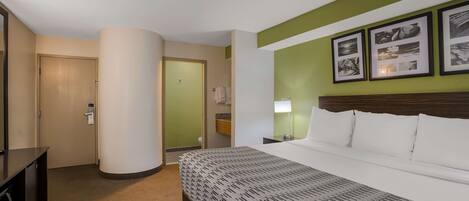 A hotel room with a bed, a desk, a chair, a lamp, and a wall with framed pictures.