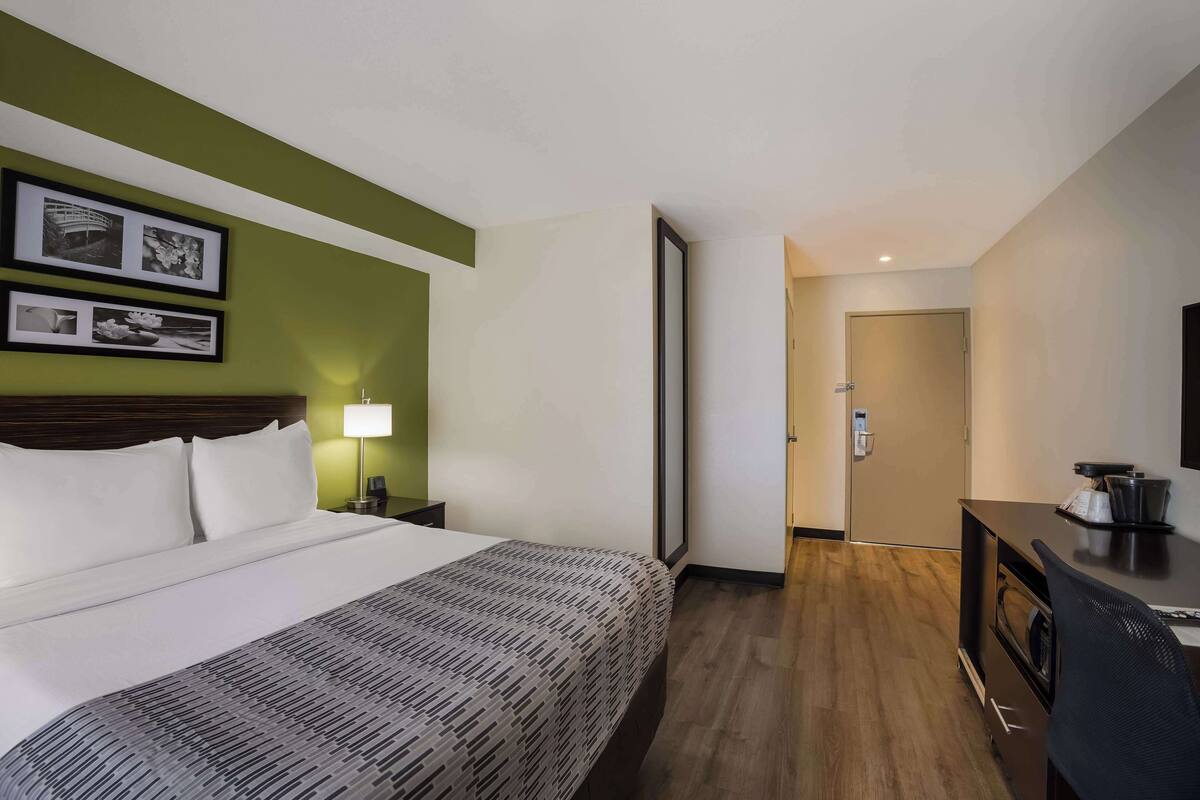 A hotel room with a bed, a desk, a lamp, and a door leading to another room.