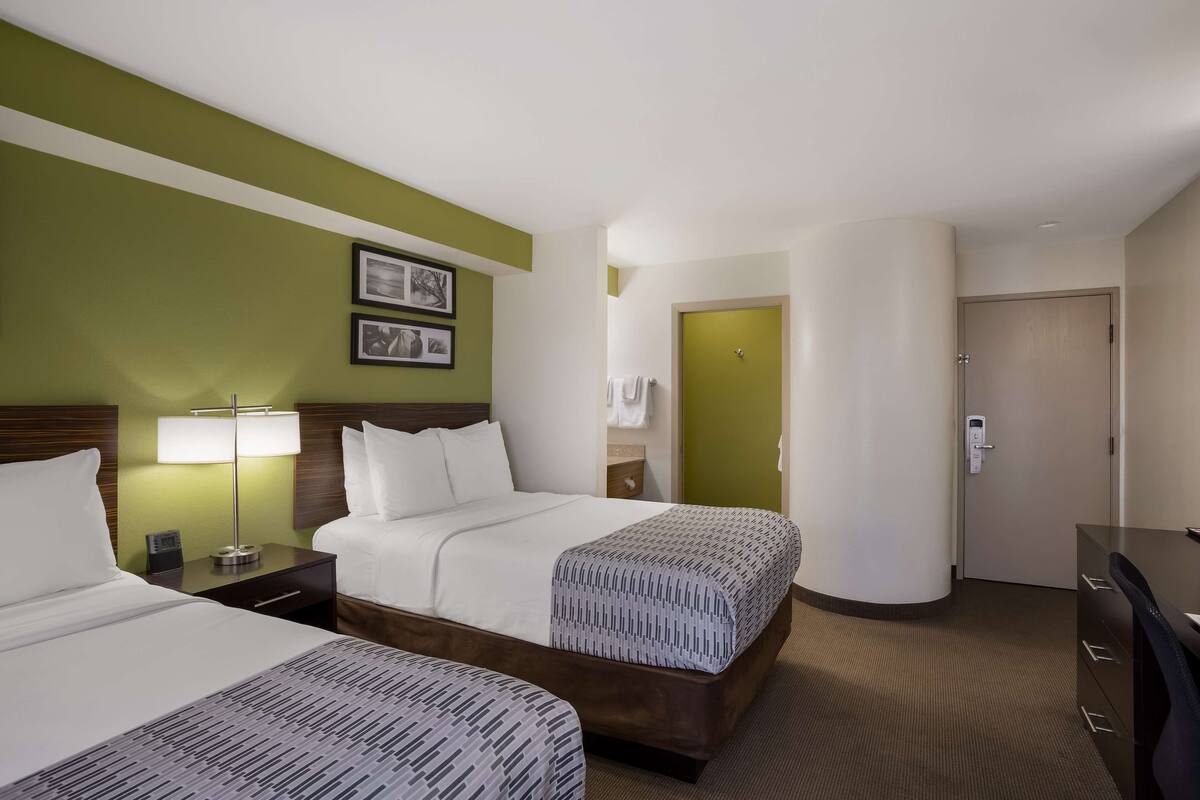 A hotel room with two beds, a nightstand with a lamp, a dresser, and a door leading to a bathroom.