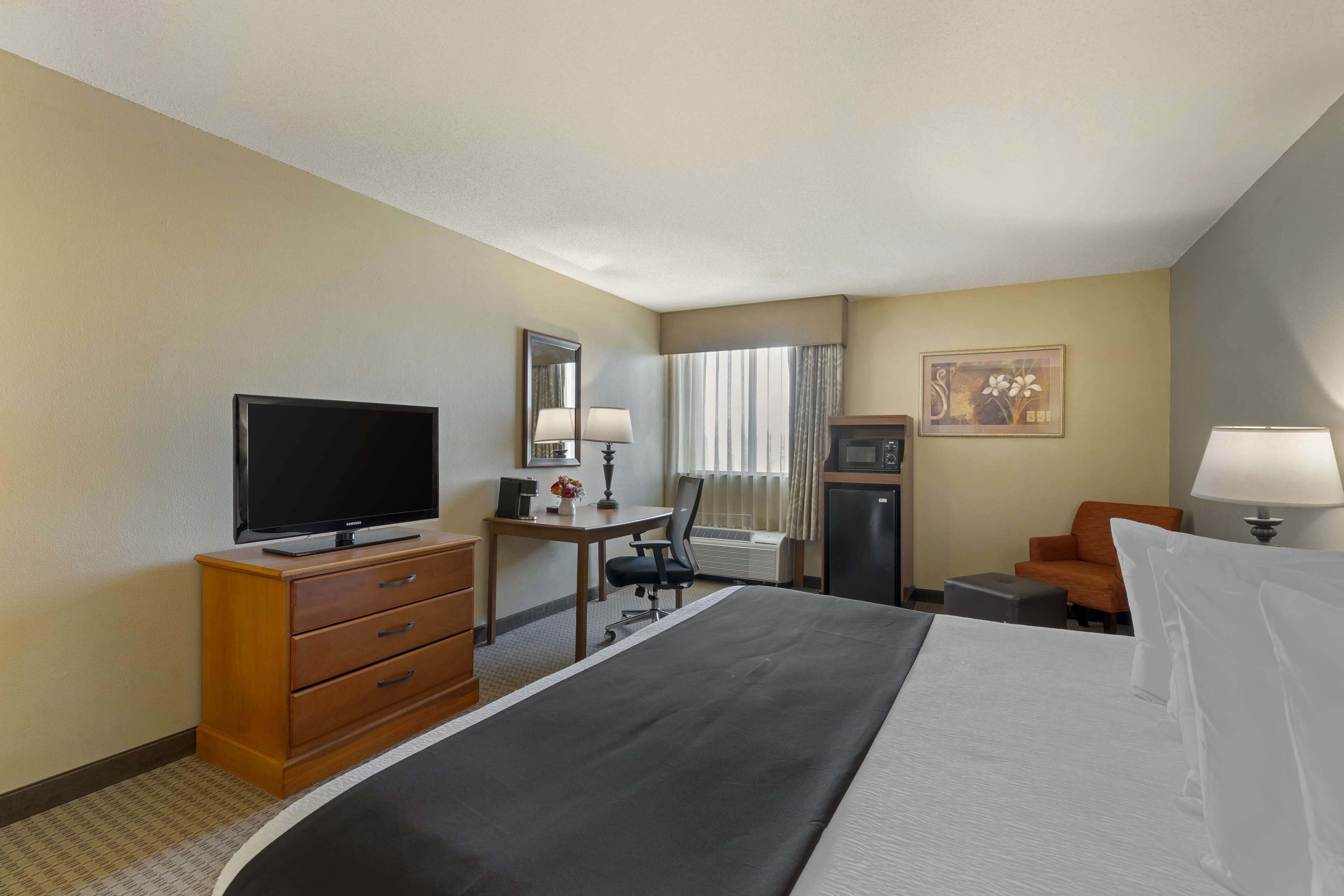 Habitación de hotel con cama, escritorio con computadora, televisor, silla, mesa pequeña y lámpara.