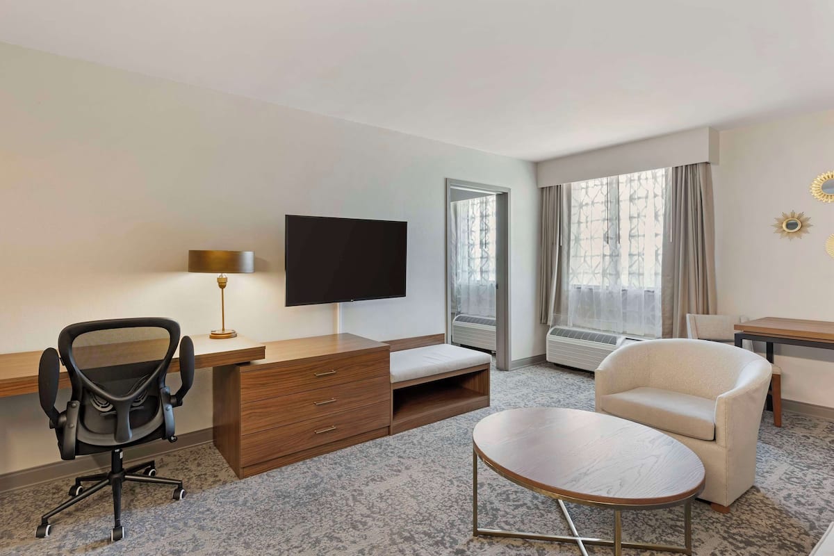 A hotel room with a desk, chair, sofa, TV, and large windows.