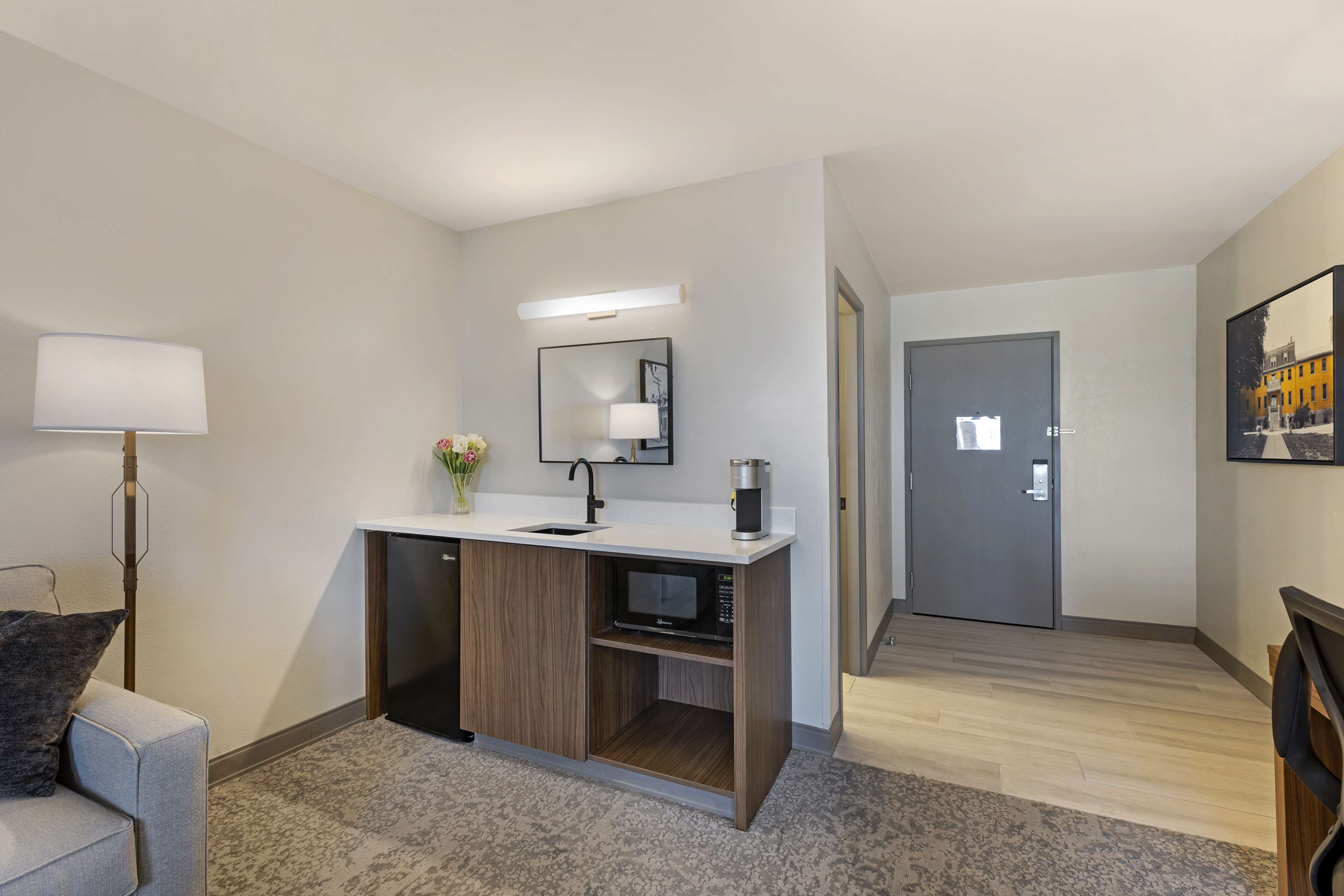 A modern hotel room with a built-in kitchenette, a large mirror, a small seating area, and a door leading to another room.