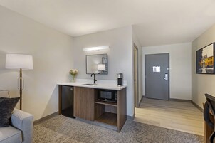 Una habitación de hotel moderna con kitchenette integrada, un espejo grande, una pequeña zona de estar y una puerta que da a otra habitación.