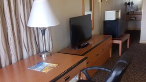 A hotel room with a desk, a television, a lamp, a chair, a microwave, and a refrigerator.