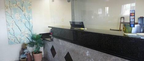 A reception desk with a glass partition, a painting on the wall, and a potted plant.