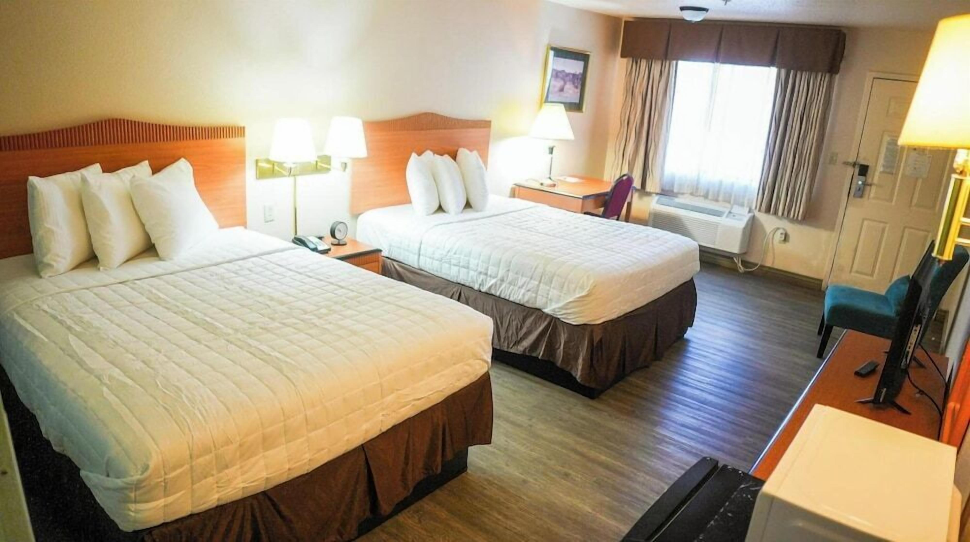 A hotel room with two beds, a desk, a chair, a window with curtains, and a wall-mounted air conditioner.