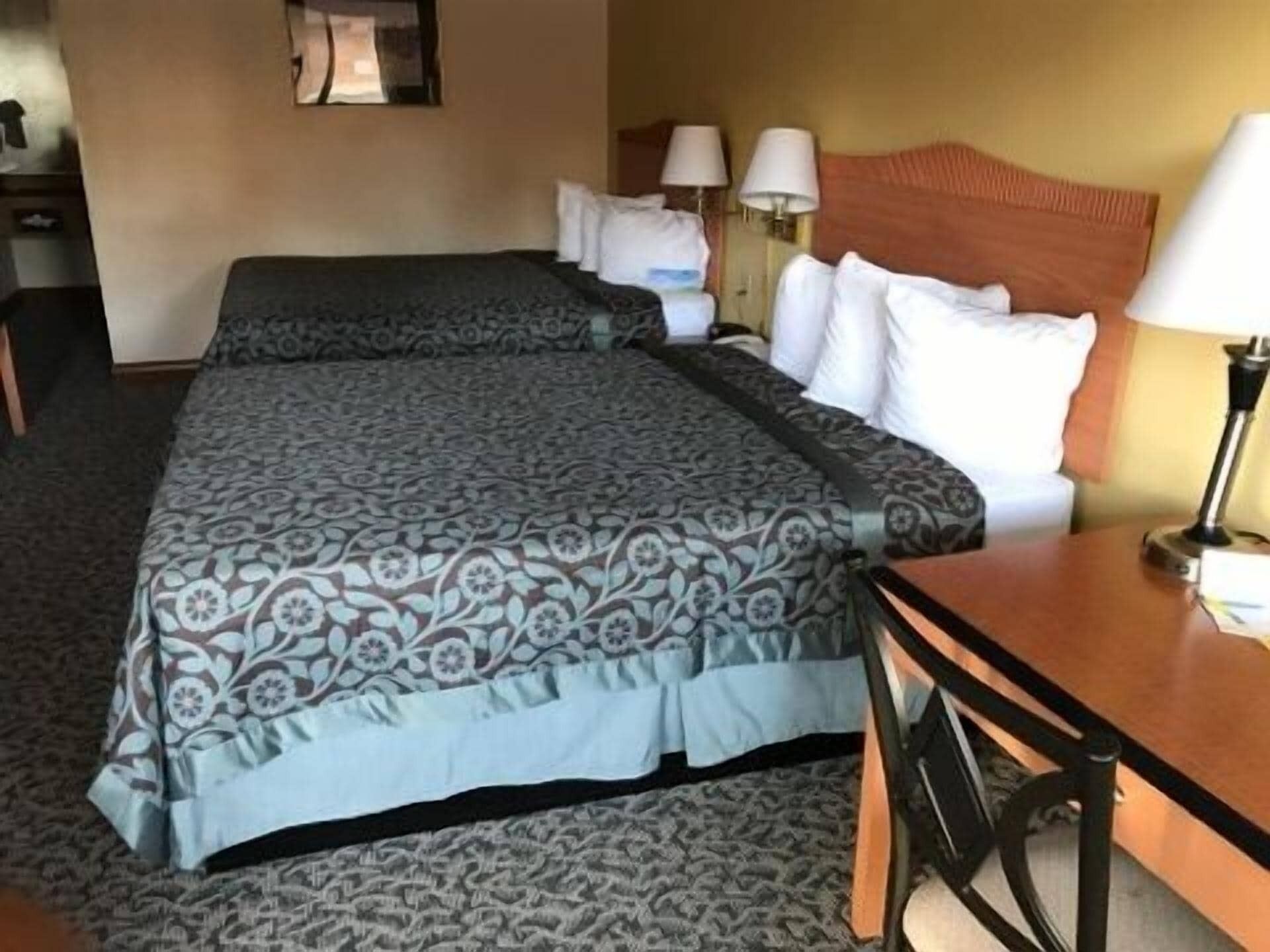 A hotel room with two beds, a wooden headboard, a desk with a chair, and two lamps.