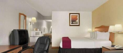 Una habitación de hotel con cama, escritorio y silla, televisor, microondas y mesita de noche con lámpara.