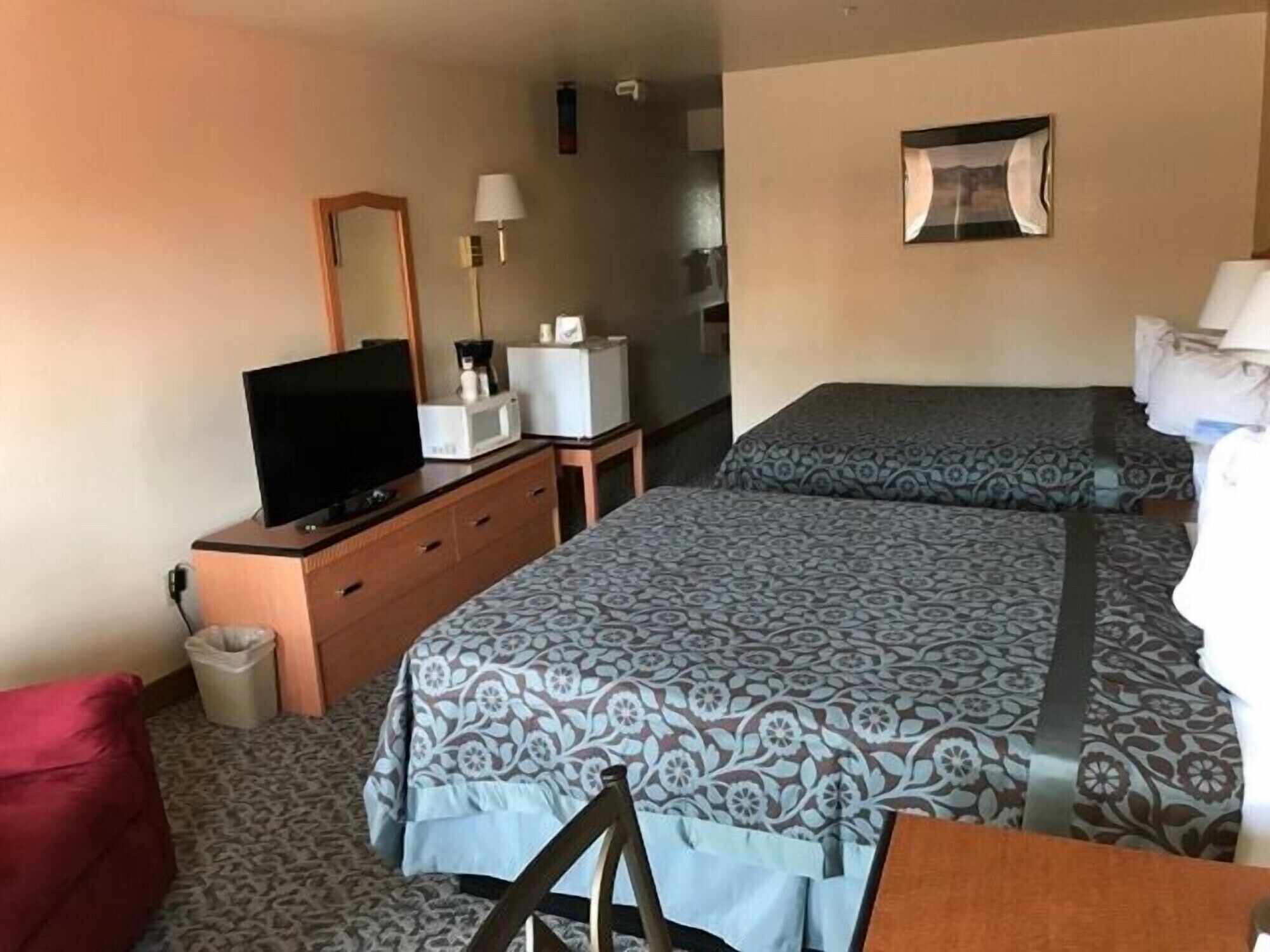 Habitación de hotel con dos camas, televisión, escritorio, silla, espejo y una mesita de noche con lámpara.