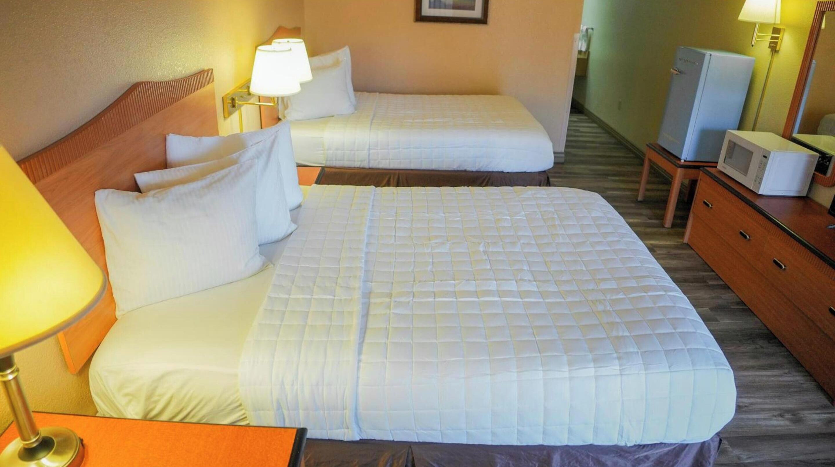 A hotel room with two beds, each with white bedding and pillows, a wooden headboard, a bedside table with a lamp, and a small TV on a wooden dresser.
