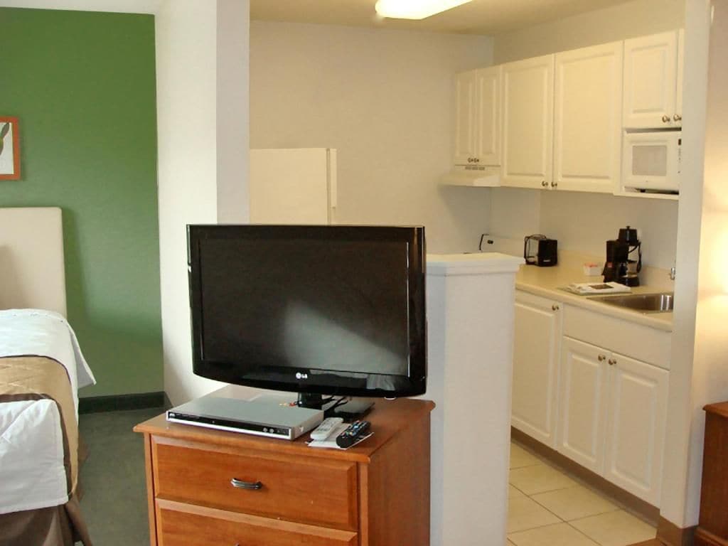 A hotel room with a bed, a television on a wooden stand, a kitchenette with white cabinets, and a microwave.