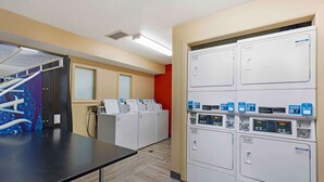 A laundry room with multiple washing machines and dryers, a drying rack, and a control panel with buttons and a display. - Studio 6 Extended Stay - St Louis, MO - Westport/Craig Road (Maryland Heights)
