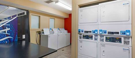 A laundry room with multiple washing machines and dryers, a drying rack, and a control panel with buttons and a display.