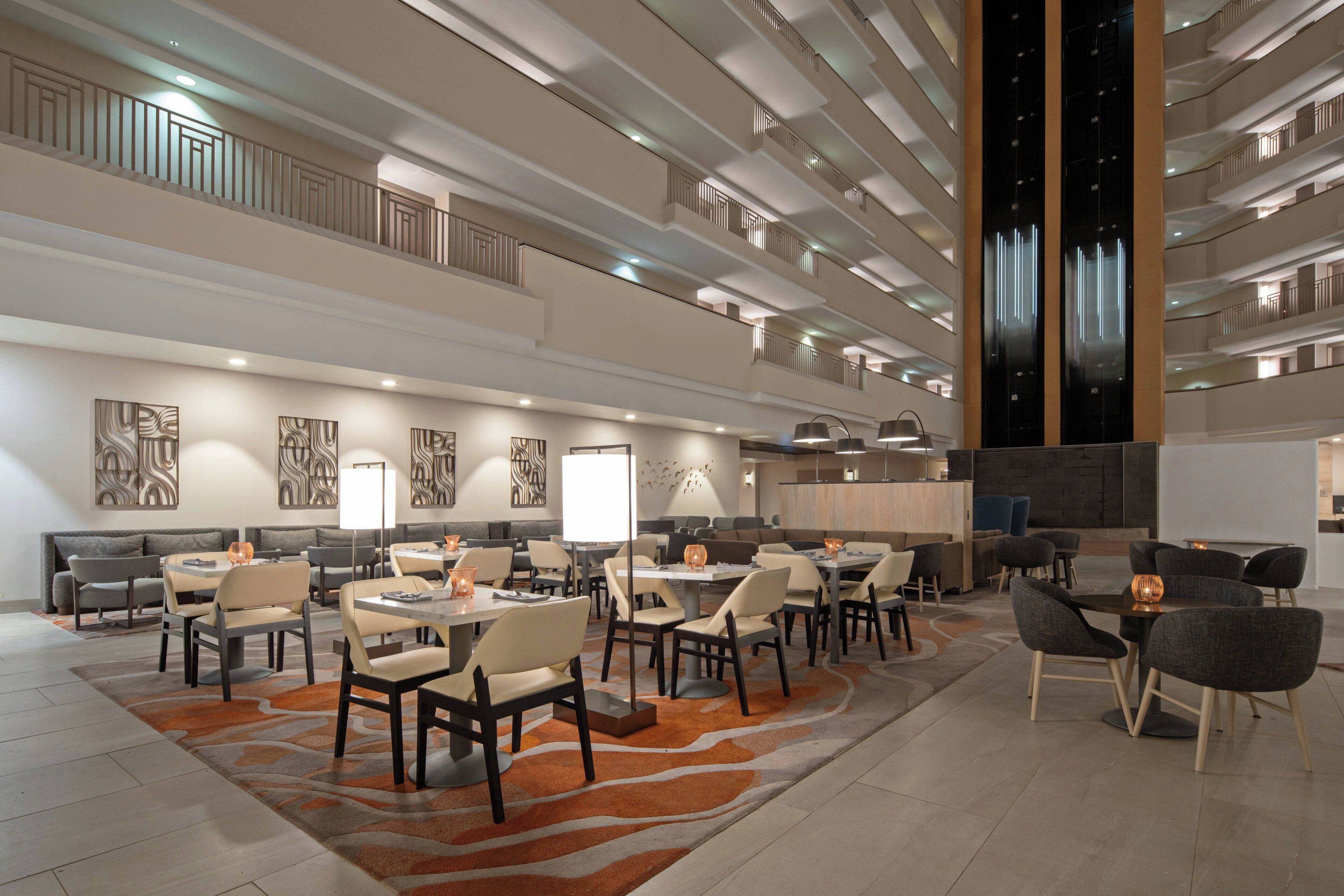 Un moderno vestíbulo de hotel con varias zonas de restauración, una recepción central y un techo alto con un elemento de iluminación único.
