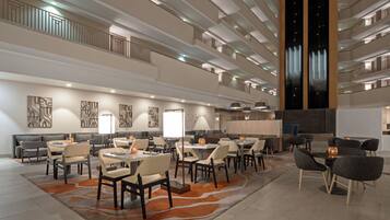 Un moderno vestíbulo de hotel con varias zonas de restauración, una recepción central y un techo alto con un elemento de iluminación único.