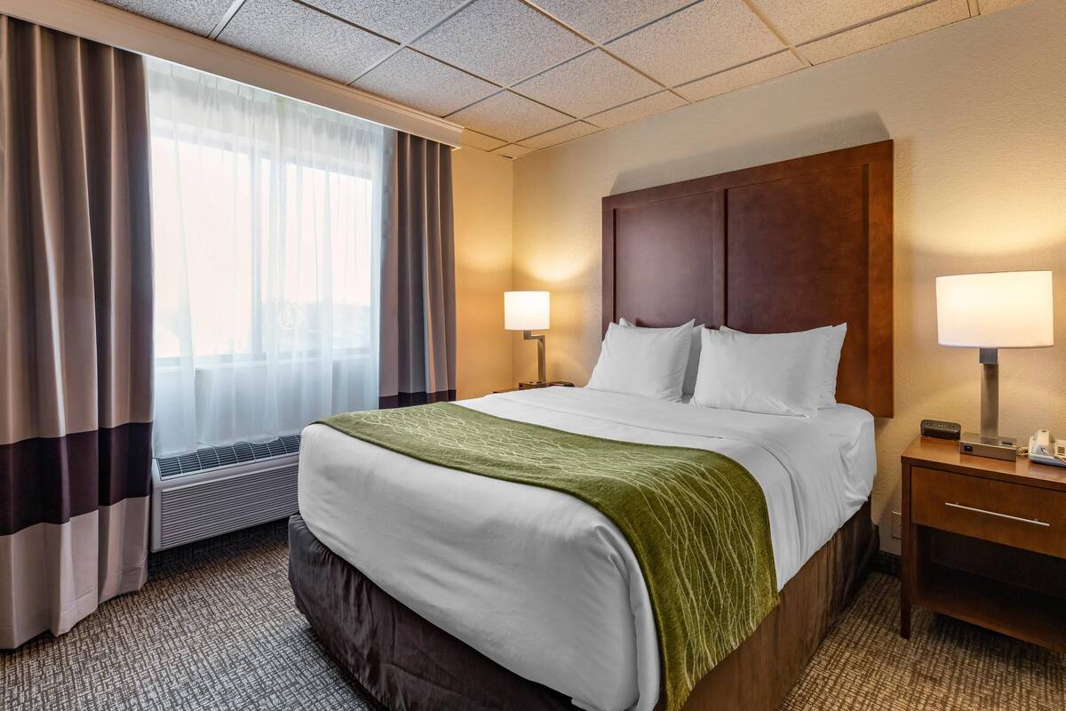 A hotel room with a large bed, a bedside table with a lamp, a window with curtains, and an air conditioning unit.
