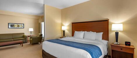 A hotel room with a large bed, a sofa, a small table, two lamps, and a nightstand with a clock.