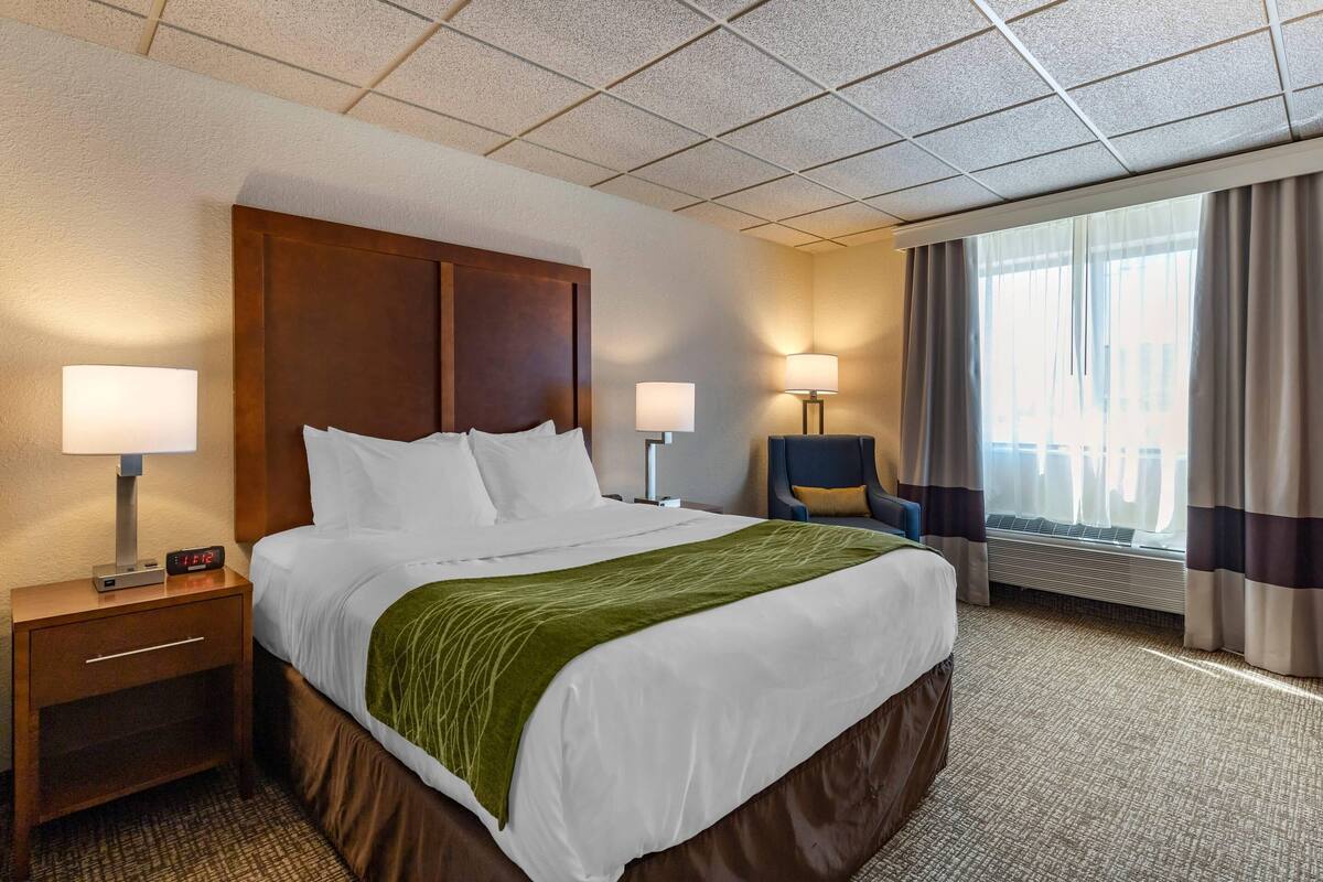 A hotel room with a large bed, two bedside lamps, a nightstand with a clock, a chair, and a window with curtains.