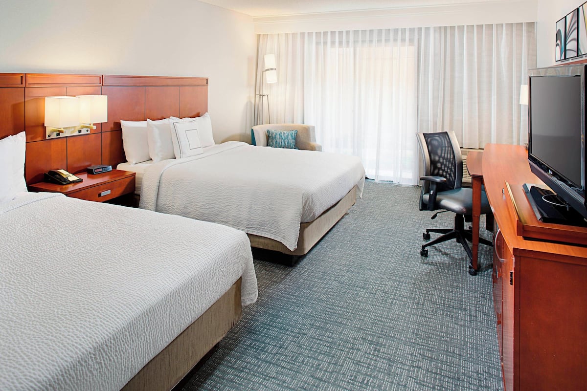Quarto de hotel com duas camas, uma escrivaninha com televisão, uma cadeira e uma mesa de cabeceira com abajur e telefone.