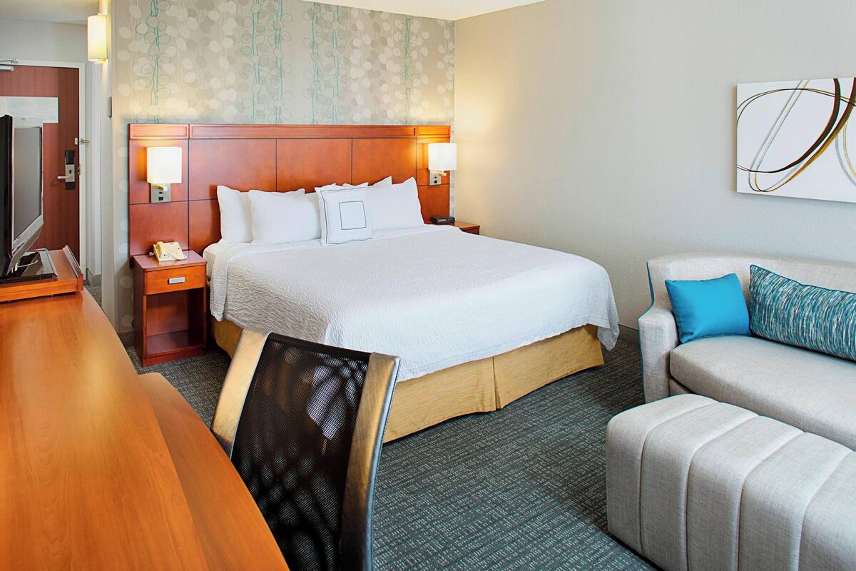 Quarto de hotel com uma cama grande, uma escrivaninha, uma cadeira, um sofá e uma televisão na parede.