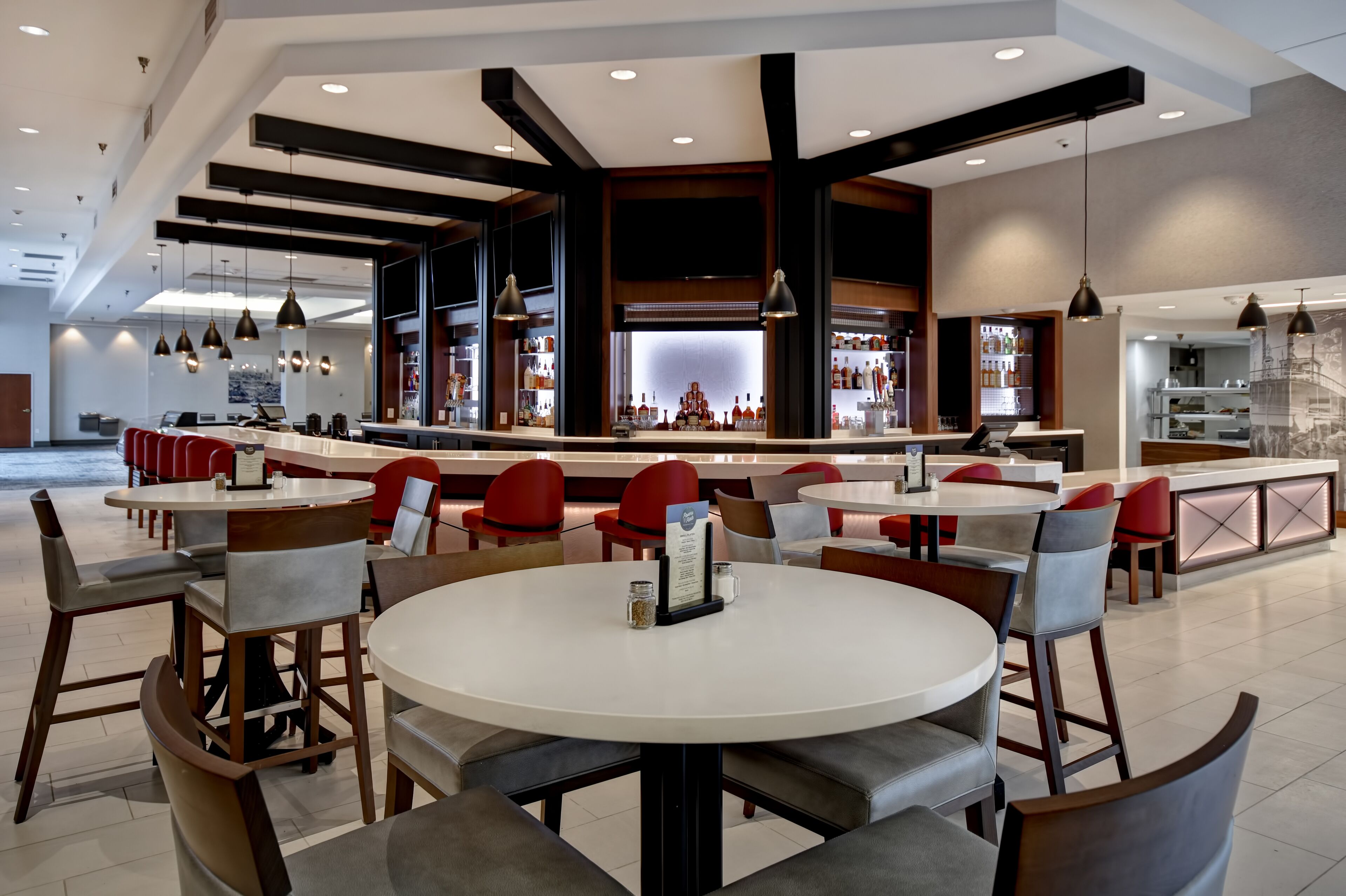 A modern restaurant with round tables and red chairs, a bar area with a counter, and pendant lighting.