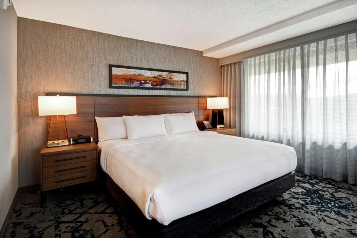 A hotel room with a large bed, two bedside tables with lamps, a painting on the wall, and a window with curtains.