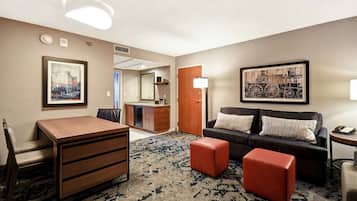 Une chambre d’hôtel avec un bureau en bois, un canapé, deux poufs rouges, un tableau représentant un paysage urbain encadré et un tableau représentant un train, également encadré.