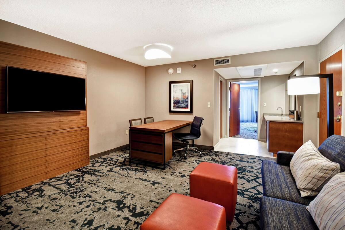A hotel room with a flat-screen TV, a wooden desk, a chair, a sofa, and a small kitchenette.
