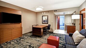 A hotel room with a flat-screen TV, a wooden desk, a chair, a sofa, and a small kitchenette.