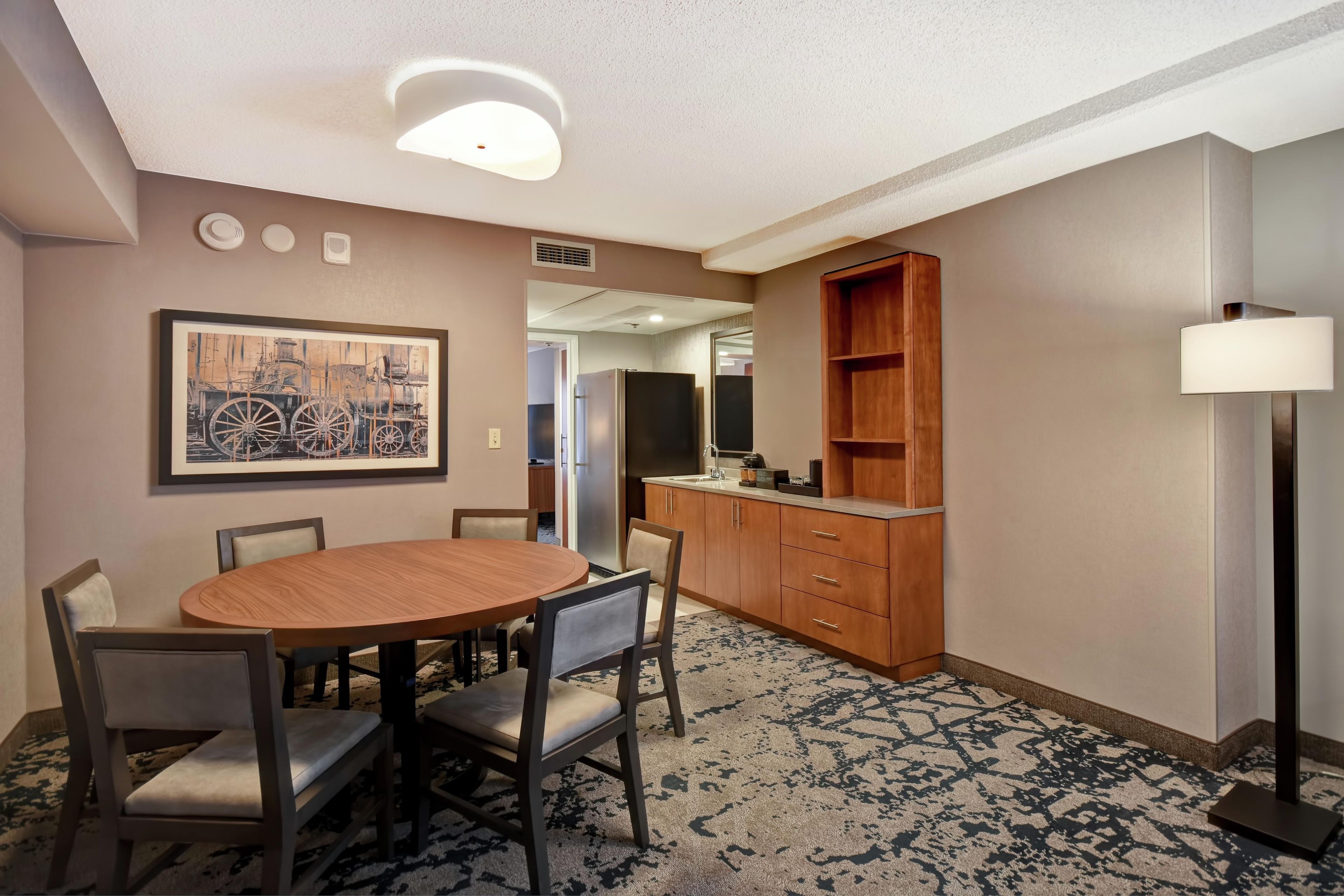 A hotel room with a dining table, chairs, a kitchen area, and a large window.