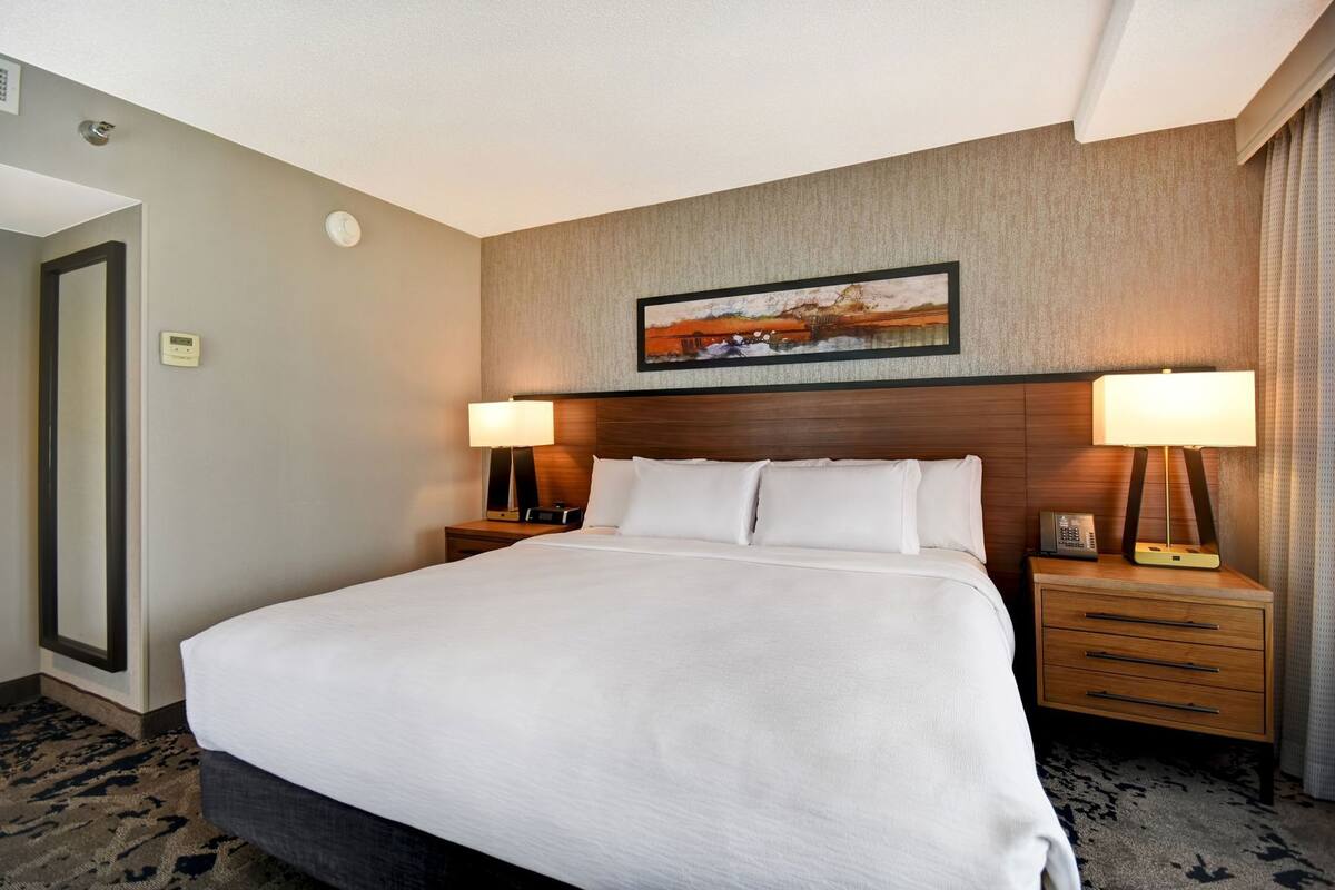 A hotel room with a large bed, two bedside tables with lamps, a painting above the bed, and a door on the left.