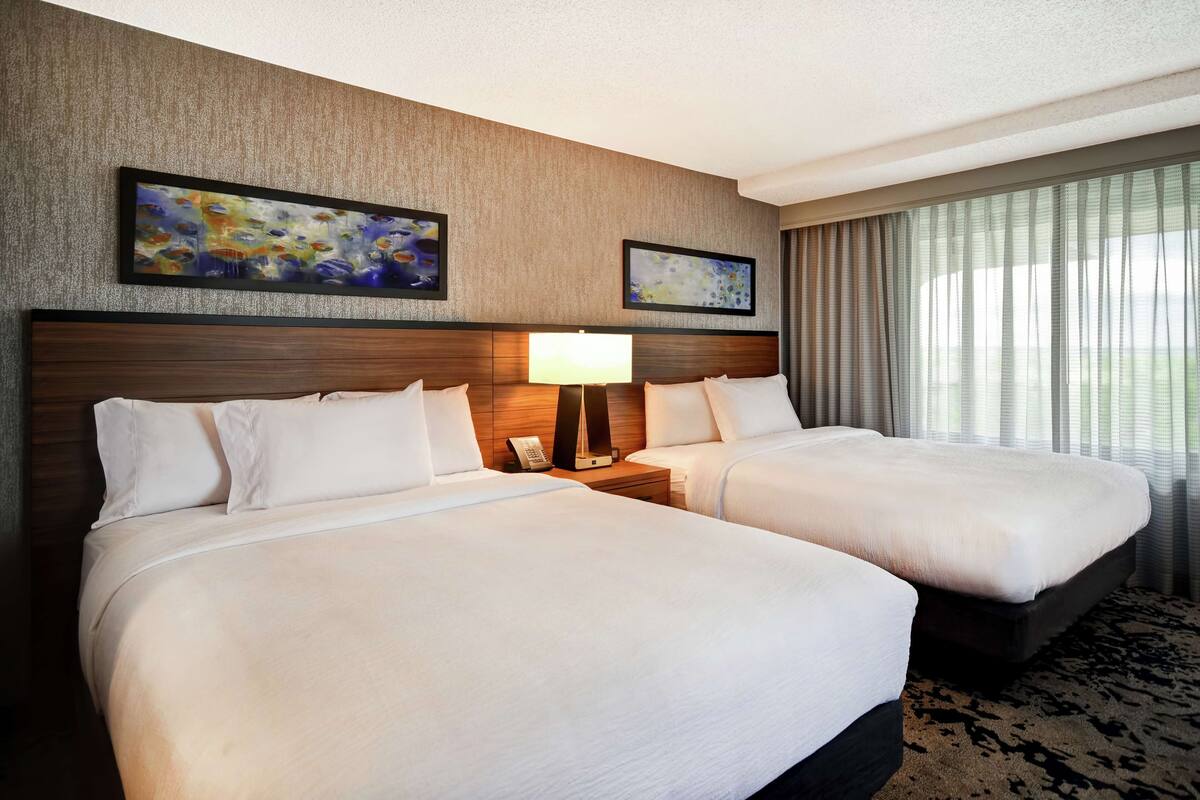 A hotel room with two beds, a wooden headboard, a bedside table with a lamp, and artwork on the wall.