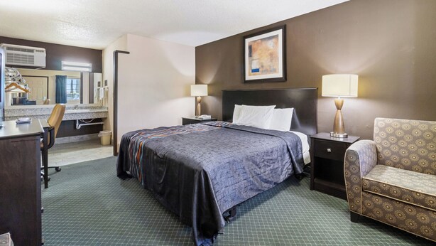 A hotel room with a bed, a desk, a chair, a sofa, a mirror, and a painting on the wall. - Economy Inn Barstow (Barstow)