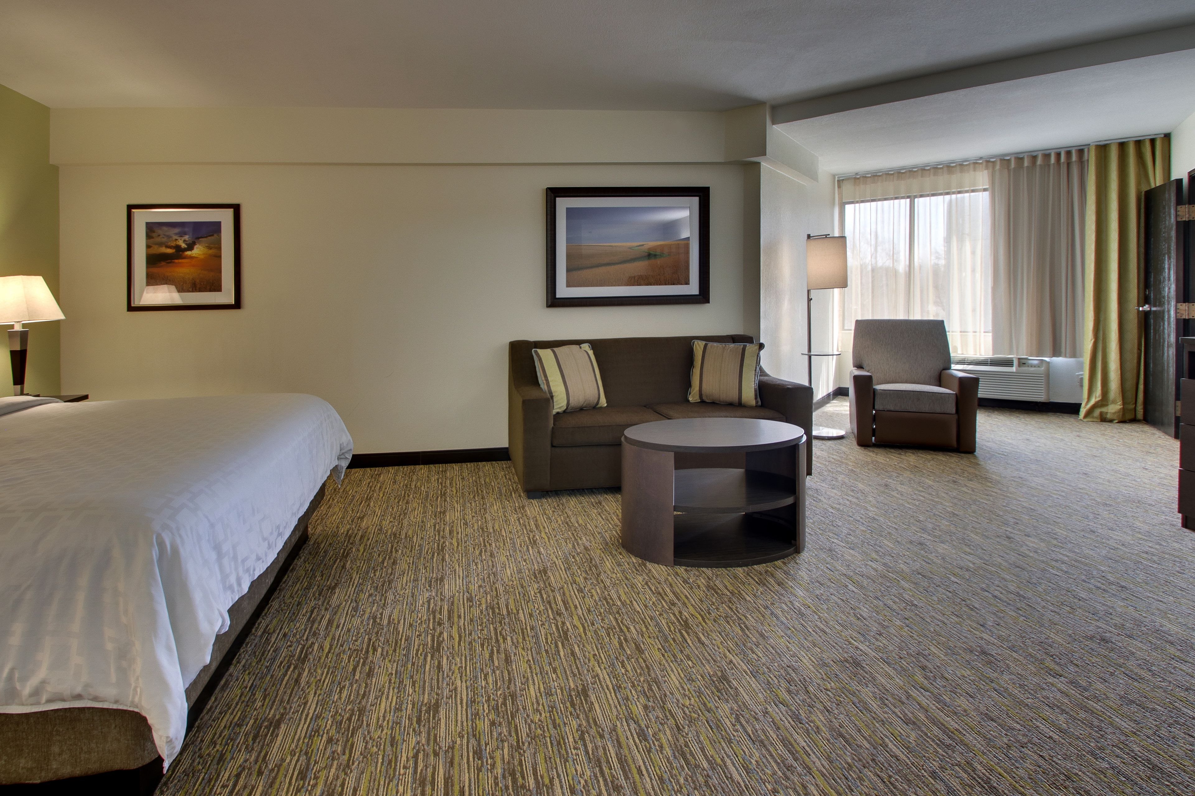 A hotel room with a bed, a sofa, a round coffee table, a chair, and two framed pictures on the wall.