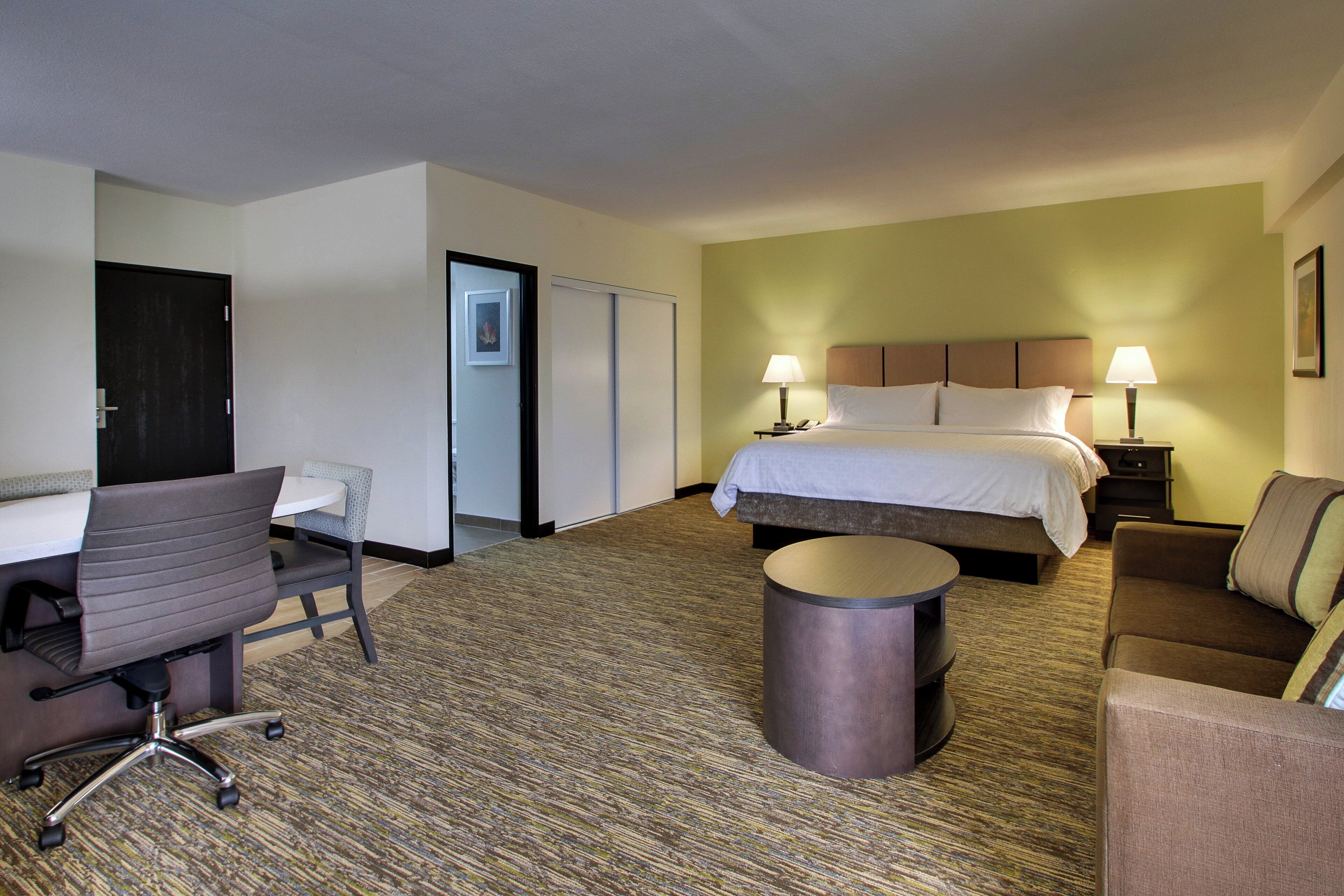 A hotel room with a bed, two bedside lamps, a desk with a chair, a round table, and a sofa.