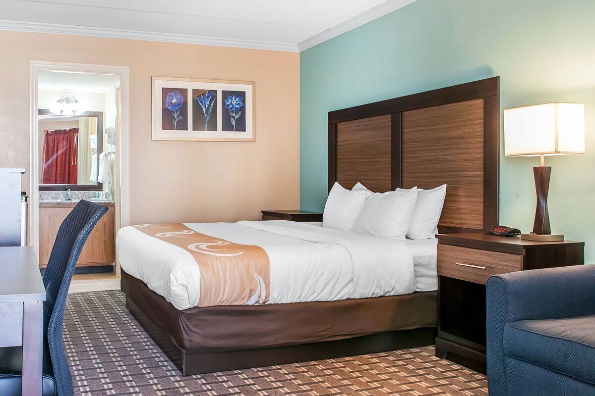 A hotel room with a bed, a desk, a chair, a nightstand, a lamp, and a wall with framed artwork.