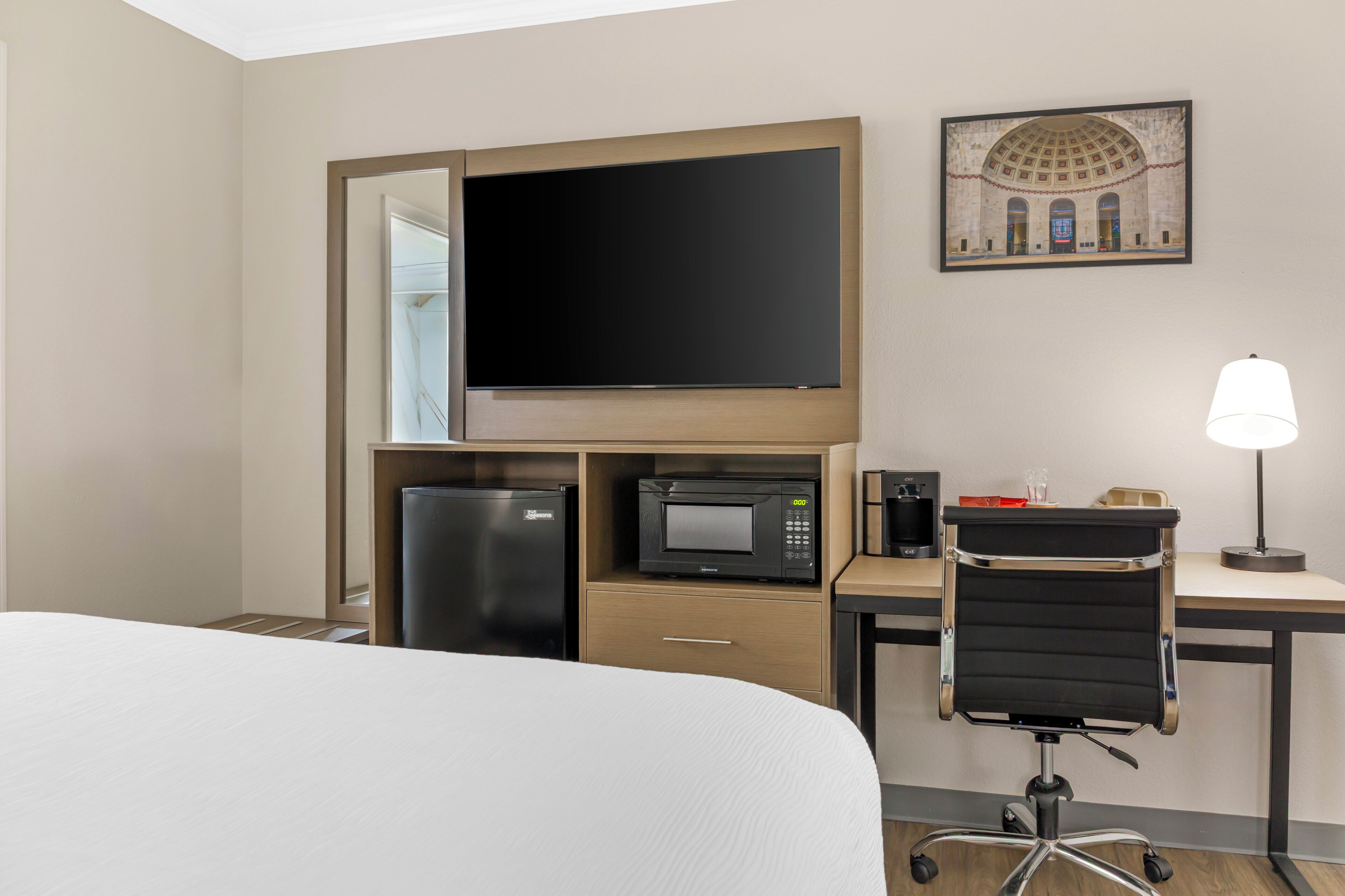 A hotel room with a bed, a desk with a computer, a microwave, a refrigerator, a chair, a lamp, and a framed picture on the wall.