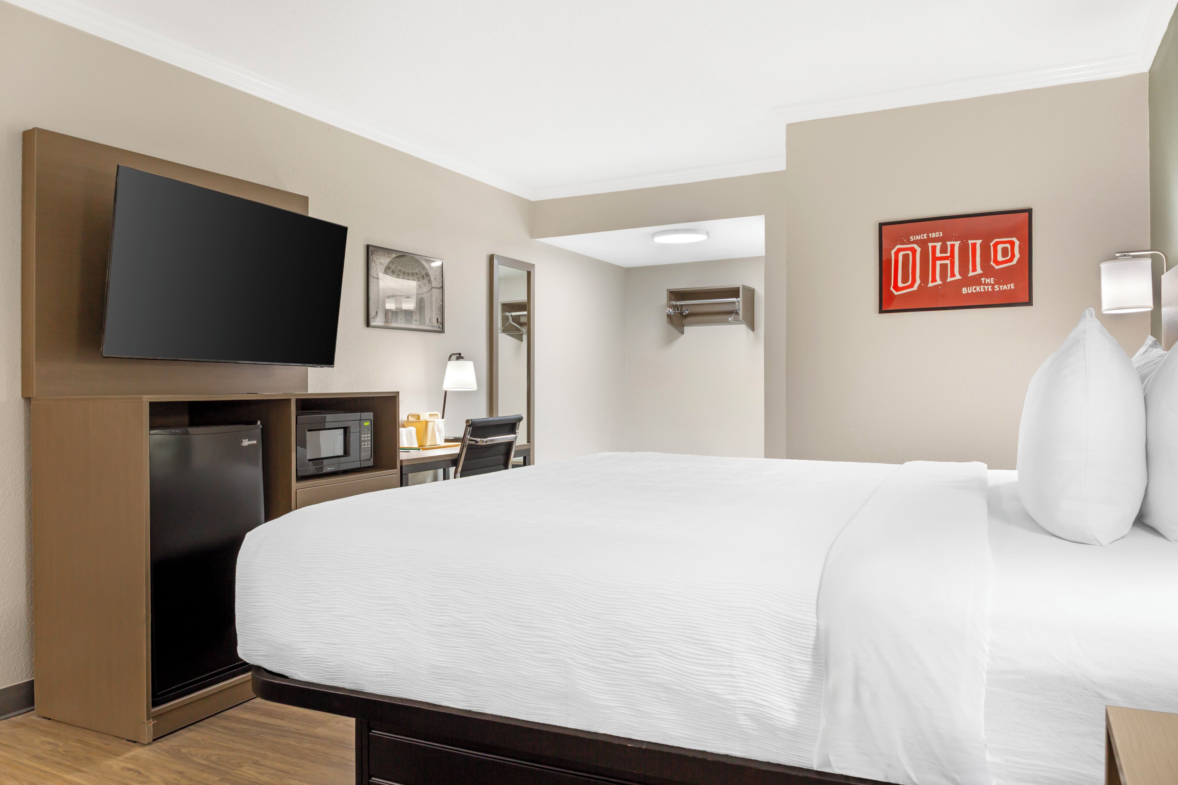 A hotel room with a bed, a flat-screen TV, a microwave, a refrigerator, a desk with a chair, and a wall-mounted air conditioning unit.