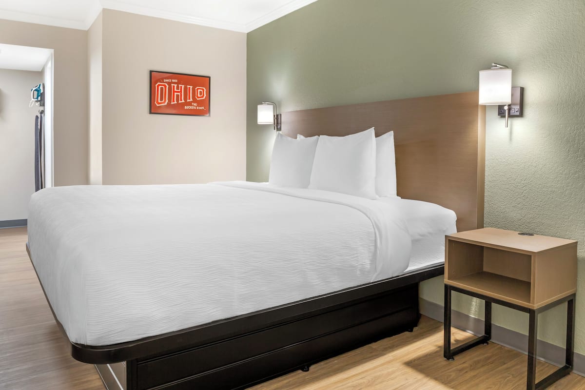 A hotel room with a large bed, a nightstand, a wall-mounted light, and a framed sign.